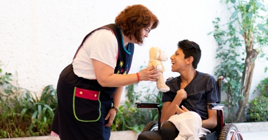 woman holding a teddy bear leaning over to talk to a child with cerebral palsy in a wheelchair