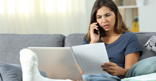 woman with an angry expression and a cast on her leg looking at a file folder and talking on the phone